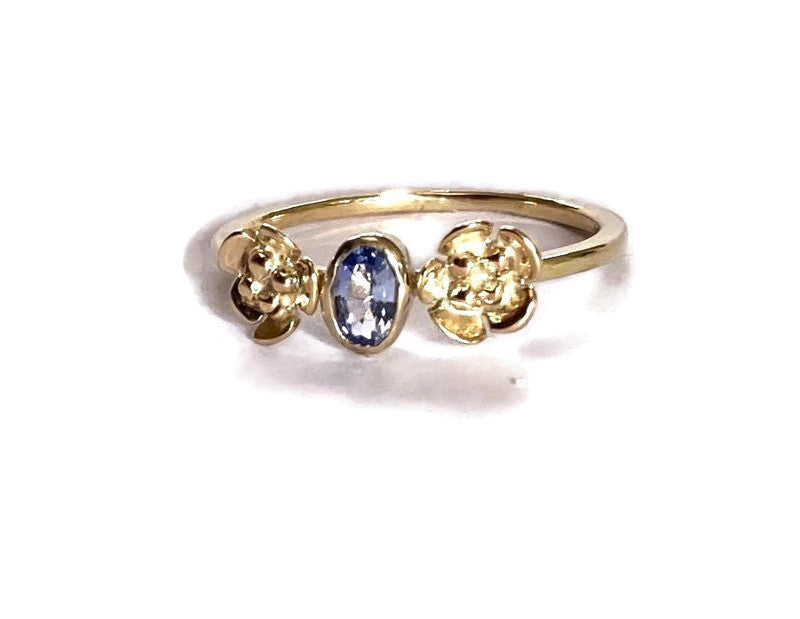 Pretty gold ring with flowers either side of central gemstone and set with light blue oval sapphire, on white background