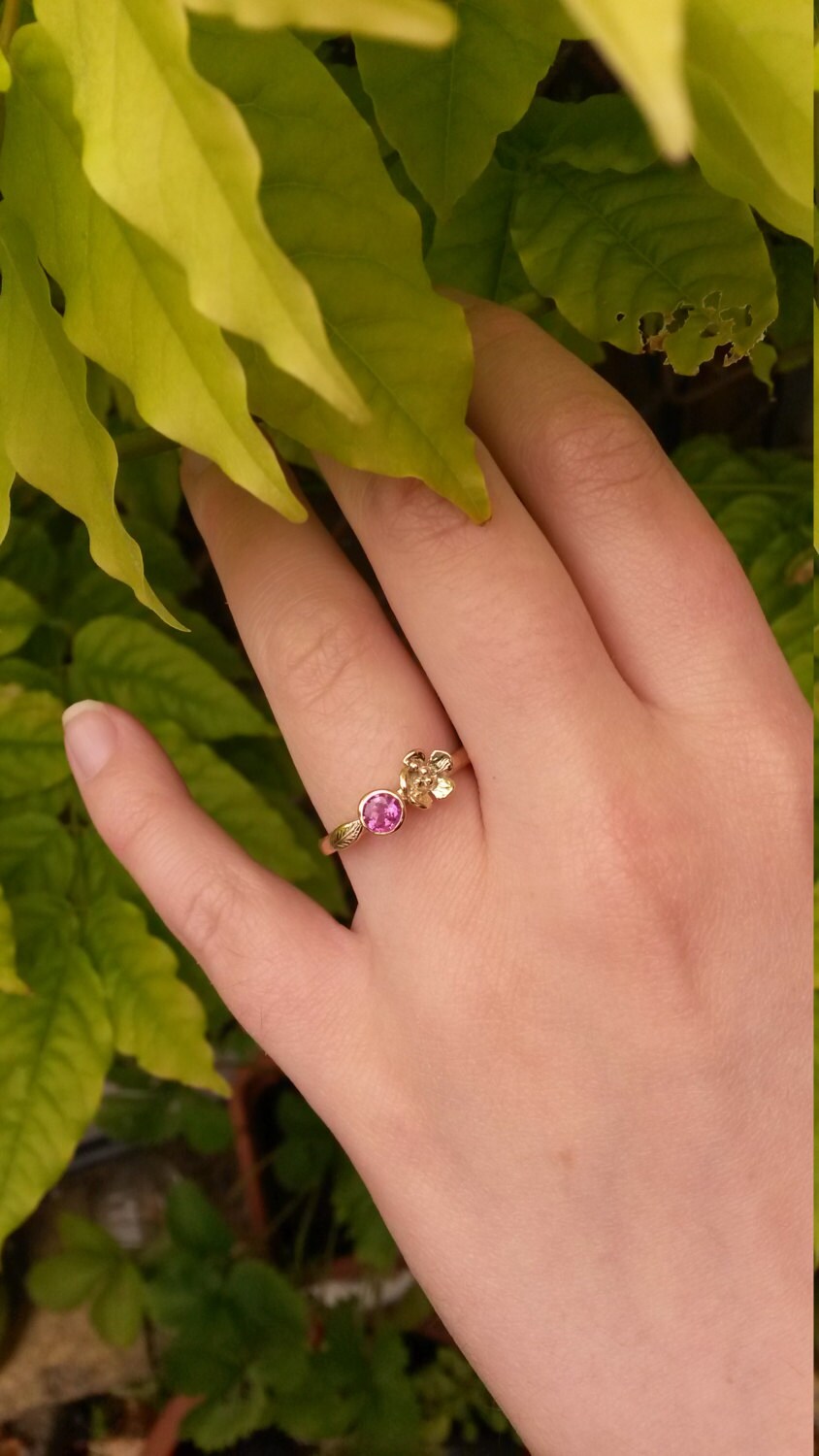 gold floral engagement ring set with vibrant pink stone and embellished with gold flower and leaf, on green background