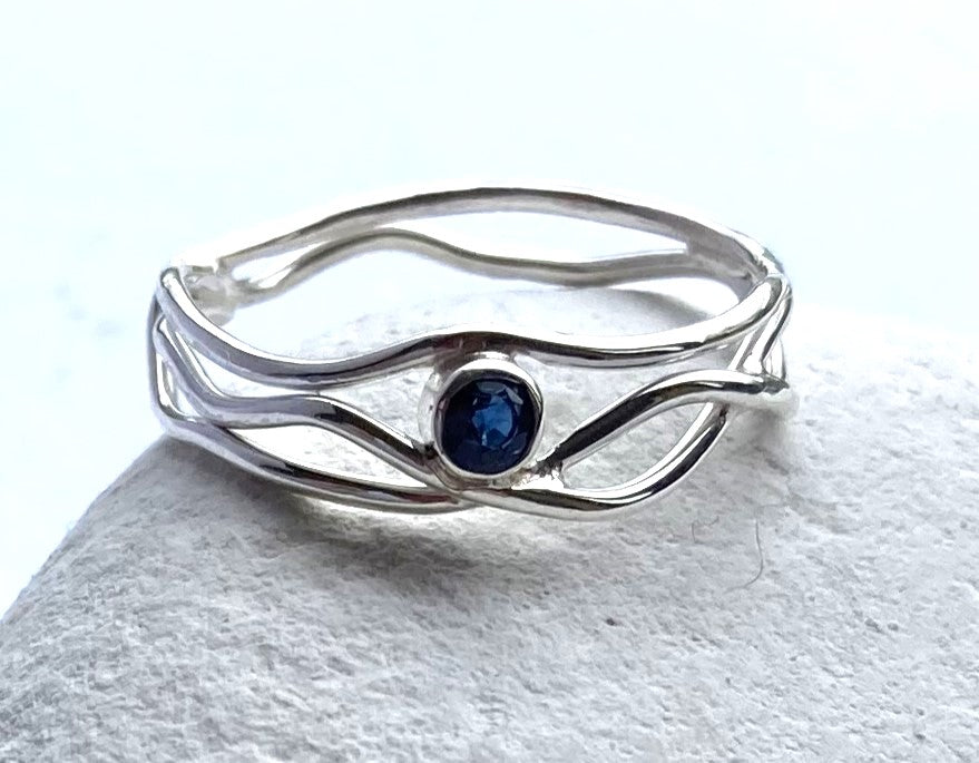 wavey silver ring set with small round blue sapphire, on white pebble