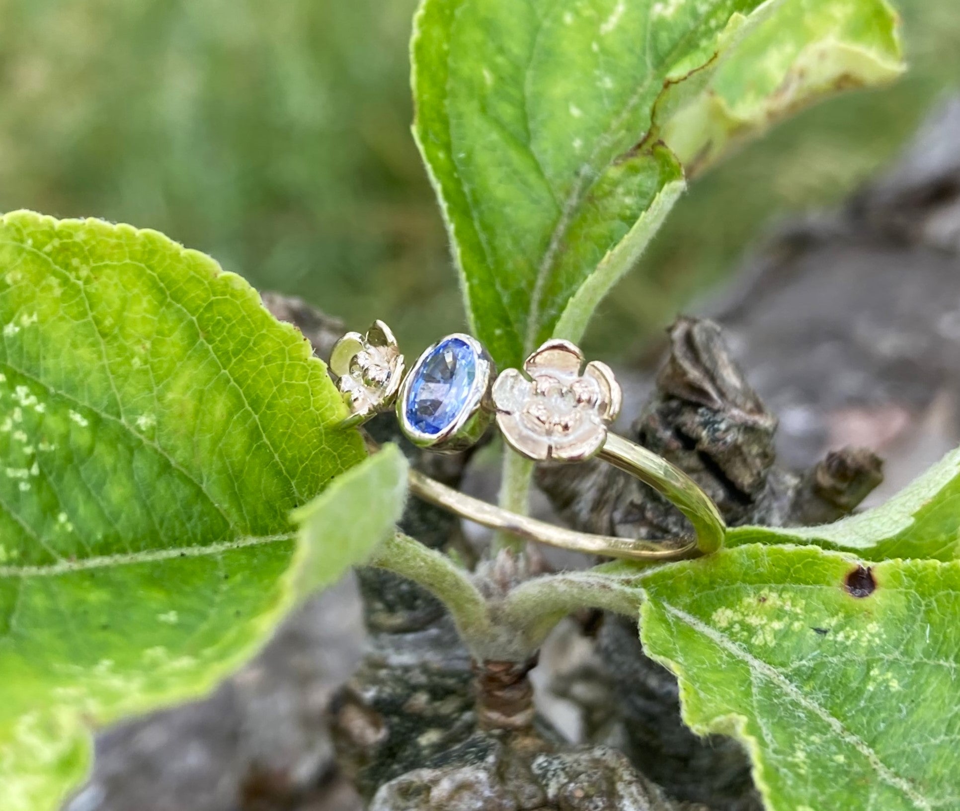 handmade gold floral ring set with pale blue oval sapphire, resting in green leaves